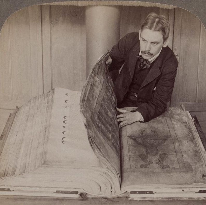 May include: A sepia-toned photograph of a man examining a large, open book. The book's pages are thick and aged, with illustrations and text visible. The man is wearing a dark suit and bow tie, engrossed in the book's contents.