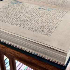 May include: An open book with aged, parchment-like pages filled with handwritten text in dark ink. The book's edges are visible, revealing the thickness of the pages. Faint blue watercolor illustrations are visible on the pages.