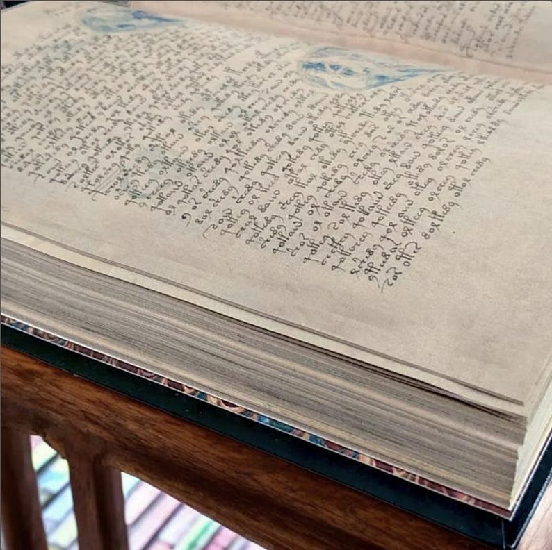 May include: An open book with aged, parchment-like pages filled with handwritten text in dark ink. The book's edges are visible, revealing the thickness of the pages. Faint blue watercolor illustrations are visible on the pages.