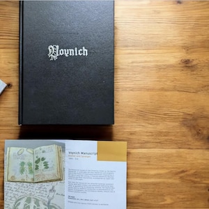 May include: A black book titled "Voynich" with white lettering, resting on a wooden surface. A brochure with the title "Voynich Manuscript" and illustrations is also present. The book and brochure are on a wooden surface.