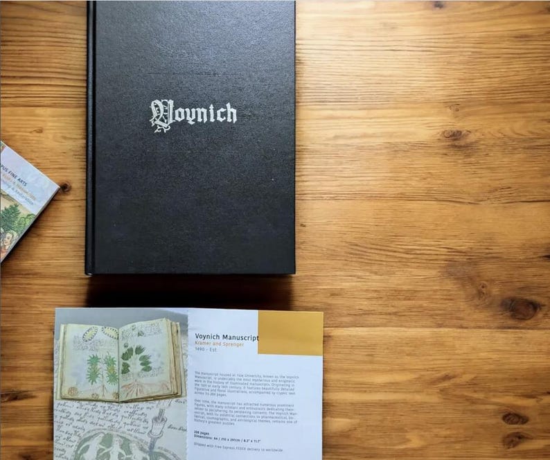 May include: A black book titled "Voynich" with white lettering, resting on a wooden surface. A brochure with the title "Voynich Manuscript" and illustrations is also present. The book and brochure are on a wooden surface.