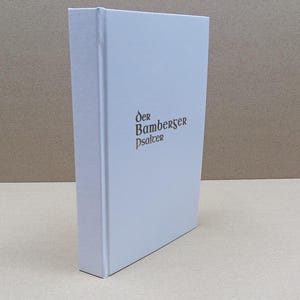 Może przedstawiać: Twarda oprawa książki z białą okładką i tytułem "Der Bamberger Psalter" w brązowych literach. Książka stoi pionowo, ukazując grzbiet i okładkę. Tło jest neutralne beżowe.