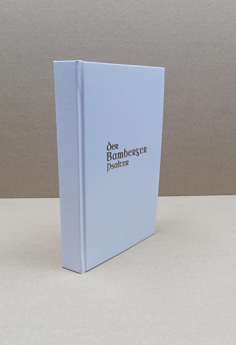Może przedstawiać: Twarda oprawa książki z białą okładką i tytułem "Der Bamberger Psalter" w brązowych literach. Książka stoi pionowo, ukazując grzbiet i okładkę. Tło jest neutralne beżowe.