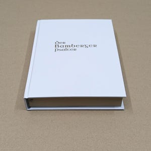 Może przedstawiać: Książka w twardej oprawie z białą okładką. Tytuł "Der Bamberger Psalter" jest wydrukowany złotą czcionką na okładce. Książka jest zamknięta i spoczywa na powierzchni w neutralnym kolorze.