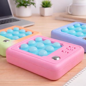 Puede incluir: Primer plano de cuatro juguetes sensoriales coloridos. Cada juguete tiene forma rectangular con una cuadrícula de burbujas azul claro. Los juguetes son de colores pastel: rosa, naranja, verde y morado. Cada juguete tiene un botón plateado.