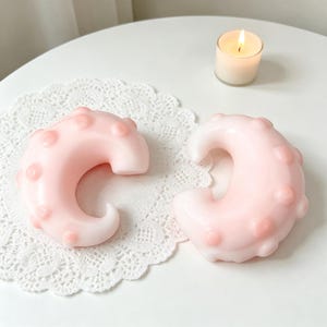 May include: Two pink, crescent-shaped candles with rounded bumps are displayed on a white lace doily. A small lit candle sits in the background. The candles have a smooth, glossy finish and a soft, pastel colour.
