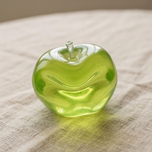 May include: A translucent green glass apple sculpture with a stem. The apple has a glossy finish and is on a neutral-coloured fabric surface. The sculpture is a decorative item.