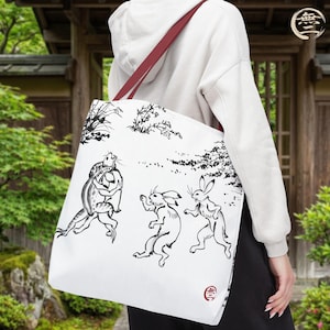 May include: A white tote bag featuring a black ink-style illustration of anthropomorphic rabbits and a frog engaged in a wrestling match. The design includes delicate botanical accents and a small red circular seal mark in the bottom corner.