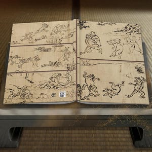 Op de afbeelding: Een open boek met zwarte inktillustraties in Chōjū-giga-stijl op een lichte achtergrond met houtnerf. De pagina's tonen antropomorfe konijnen en kikkers die verschillende activiteiten uitvoeren, zoals worstelen, rennen en dragen.