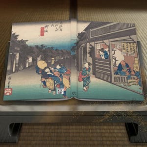 以下が含まれることがあります： 日本の浮世絵のイラストが描かれた開いた本。伝統的な衣装を着た人物、建物、青い空が描かれた街並みの版画です。本は濃い灰色のスタンドに展示されています。