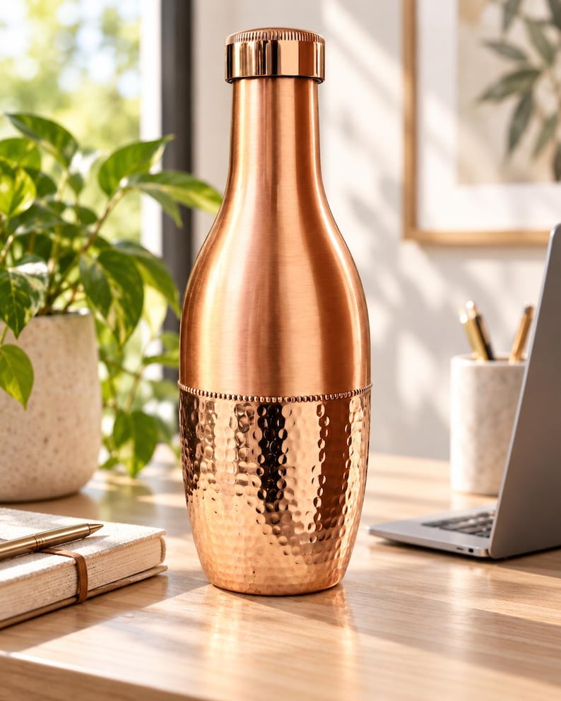 May include: A copper-colored metal bottle with a smooth, polished upper section and a textured, hammered lower section. The bottle features a cylindrical neck, a rounded body, and a matching metallic cap that seals the top of the vessel securely.