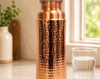 May include: A cylindrical copper water bottle with a hammered texture on the main body and a smooth neck and cap. The bottle features a metallic finish and is branded with the text SSA SHIV SHAKTI ARTS in a yellow oval at the top left corner.