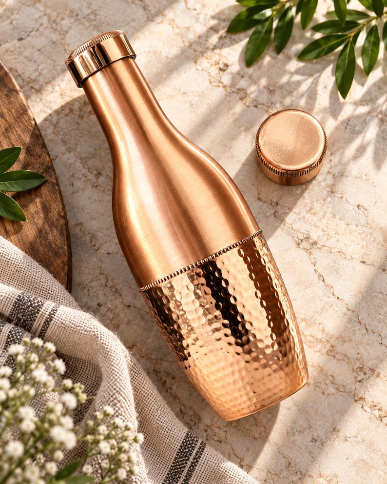 May include: A copper water bottle with a smooth upper neck and a textured, hammered lower body. The bottle features a cylindrical screw-top lid and a separate matching circular cap resting nearby, showcasing a metallic finish and reflective surface.