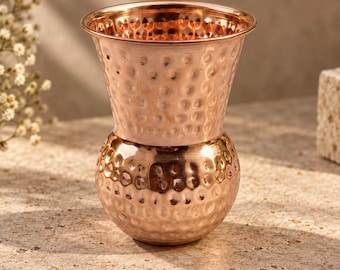 May include: A polished copper vessel featuring a distinct hammered texture across its entire surface. The object has a rounded, bulbous base that tapers inward before widening into a flared, open rim, creating a classic, symmetrical silhouette.