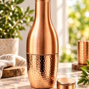 May include: A copper water bottle with a smooth upper neck and a textured, hammered finish on the lower half. The bottle features a cylindrical shape with a narrow opening and a separate, matching flat copper cap resting beside the base.