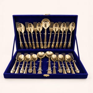 May include: A gold-toned cutlery set arranged in a blue velvet-lined storage case. The set includes multiple forks and spoons with ornate detailing on the handles, secured in place by white elastic loops within the open display box.