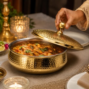 May include: A round, gold-colored metal serving bowl with a hammered texture and a matching lid featuring a spherical knob. The bowl contains a cooked dish with chunks of food in a reddish-orange sauce garnished with green herbs.