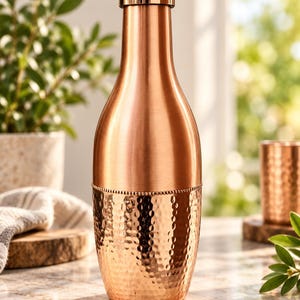 Puede incluir: Una botella de agua de cobre con una sección superior lisa y una sección inferior martillada. La botella presenta un cuerpo cilíndrico que se estrecha hacia el cuello y está rematada con una tapa metálica a juego. El acabado es de cobre pulido.