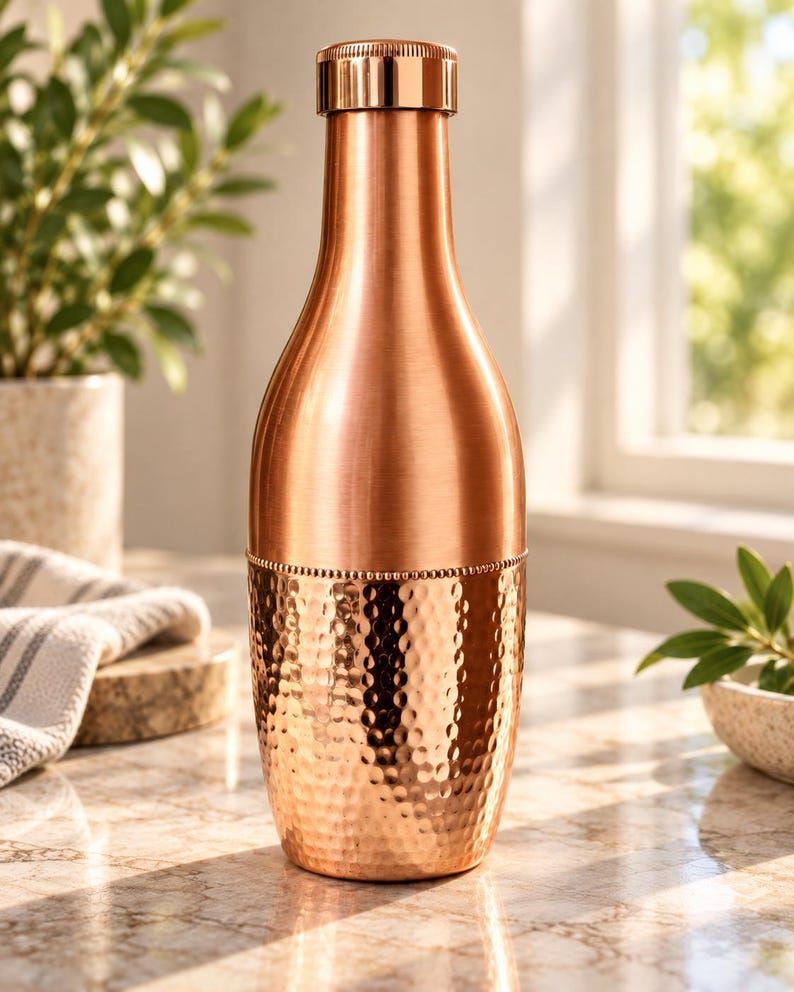 May include: A copper-colored metal water bottle with a smooth upper section and a hammered texture on the lower half. The bottle features a cylindrical shape, a narrow neck, and a matching metallic screw-top lid with a beaded detail at the center.
