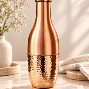 May include: A copper-colored metal water bottle with a smooth upper section and a textured, hammered lower section. The bottle features a cylindrical body that tapers toward the neck and is topped with a matching metallic screw-top cap.
