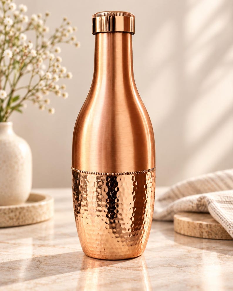 May include: A copper-colored metal water bottle with a smooth upper section and a textured, hammered lower section. The bottle features a cylindrical body that tapers toward the neck and is topped with a matching metallic screw-top cap.