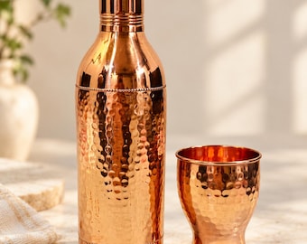 May include: A polished copper water bottle and a matching copper cup. The bottle features a tall, slender neck with a textured, hammered finish on the main body, accented by beaded detailing at the top and bottom. The cup has a matching hammered texture.