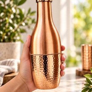 May include: A copper water bottle with a smooth upper section and a hammered lower section featuring a beaded texture band at the center. The bottle has a cylindrical neck and a matching metallic screw-top cap with a polished finish.