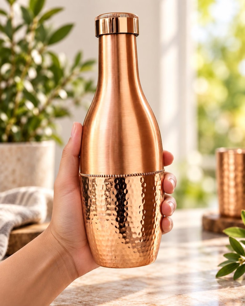 May include: A copper water bottle with a smooth upper section and a hammered lower section featuring a beaded texture band at the center. The bottle has a cylindrical neck and a matching metallic screw-top cap with a polished finish.