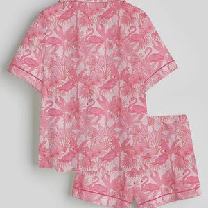 K&ouml;nnte beinhalten: Rosa Pyjama-Set mit Flamingo- und Tropenpflanzenmuster. Das kurz&auml;rmelige Oberteil und die Shorts haben einen hellrosa Hintergrund mit einem sich wiederholenden Design aus rosa Flamingos und Bl&auml;ttern. Die Kanten sind mit einer dunkleren rosa Paspel versehen.