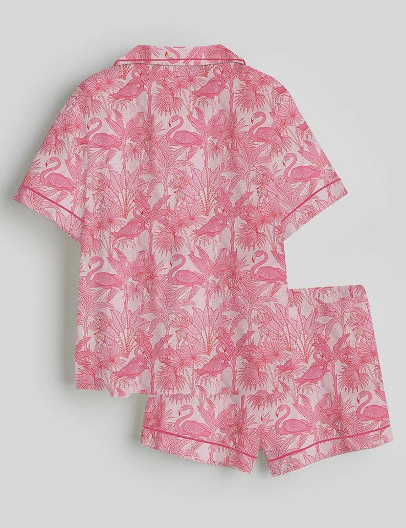 K&ouml;nnte beinhalten: Rosa Pyjama-Set mit Flamingo- und Tropenpflanzenmuster. Das kurz&auml;rmelige Oberteil und die Shorts haben einen hellrosa Hintergrund mit einem sich wiederholenden Design aus rosa Flamingos und Bl&auml;ttern. Die Kanten sind mit einer dunkleren rosa Paspel versehen.
