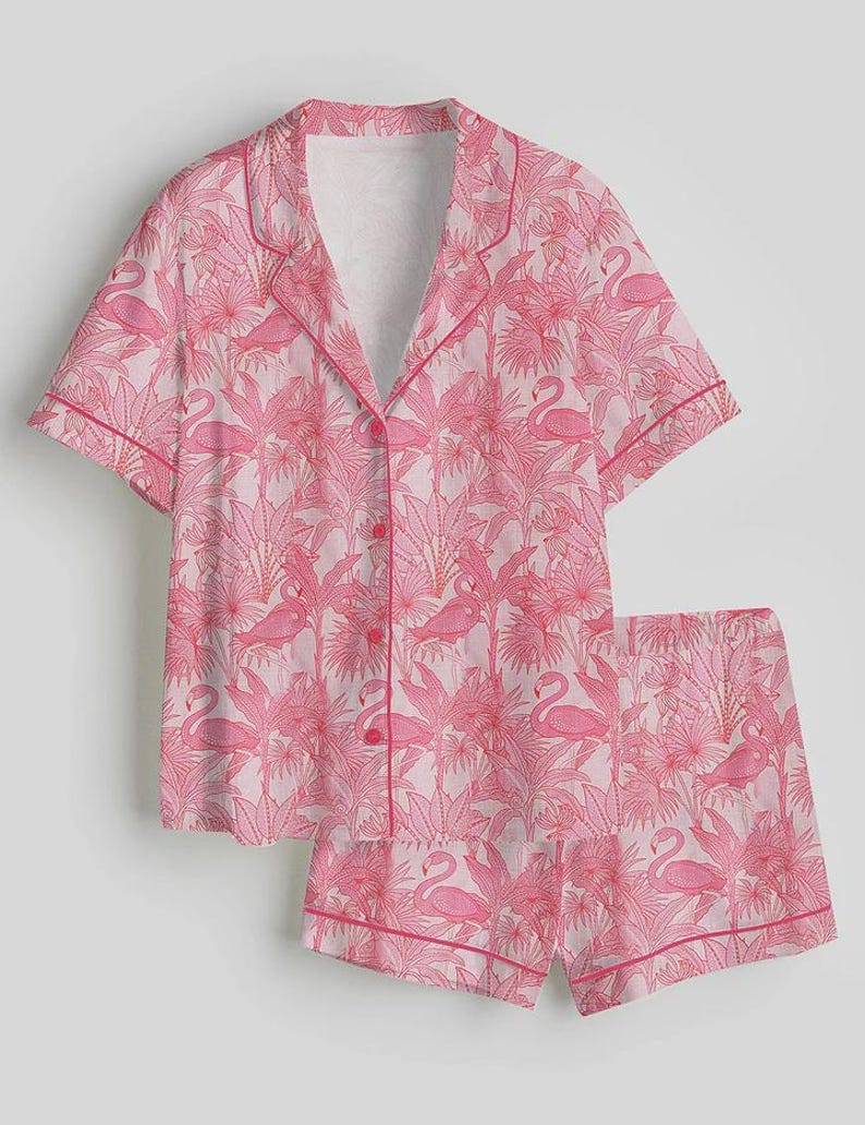 K&ouml;nnte beinhalten: Rosa Pyjama-Set mit Flamingo- und tropischem Bl&auml;ttermuster. Das kurz&auml;rmelige Hemd mit Kn&ouml;pfen und die passenden Shorts haben eine rosa Paspelierung. Der Stoff hat einen hellrosa Hintergrund mit einem sich wiederholenden Design aus rosa Flamingos und Bl&auml;ttern.