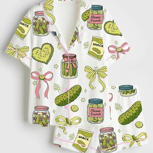 K&ouml;nnte beinhalten: Wei&szlig;es Pyjama-Set mit einem Gurkenmuster. Das Set besteht aus einem kurz&auml;rmeligen Hemd mit Kn&ouml;pfen und Shorts. Das Muster zeigt Gl&auml;ser mit Gurken, Gurkenscheiben, Schleifen und Snack-T&uuml;ten. Die Einfassung ist rosa.