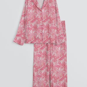 K&ouml;nnte beinhalten: Ein rosa Pyjama-Set mit einem Flamingo- und tropischen Pflanzenmuster. Das Set besteht aus einem Hemd mit Kn&ouml;pfen, einem Reverskragen und langen &Auml;rmeln sowie einer passenden Hose. Die Kanten des Hemdes und der Hose sind mit einer dunkleren rosa Paspel versehen.