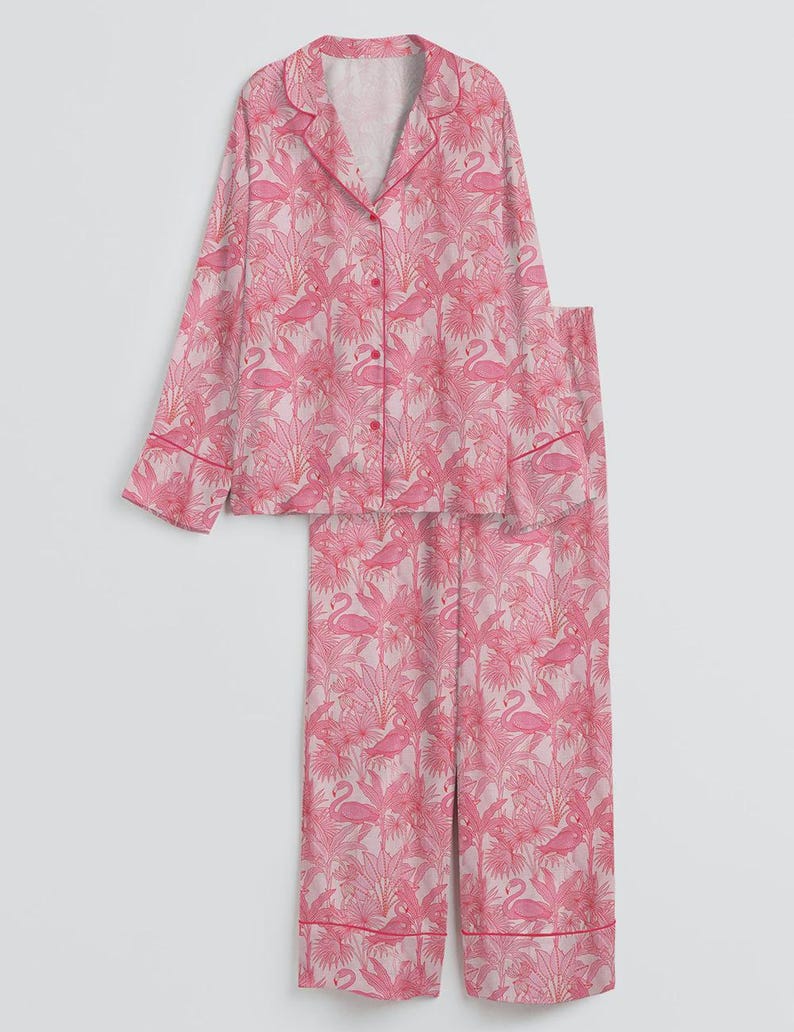 K&ouml;nnte beinhalten: Ein rosa Pyjama-Set mit einem Flamingo- und tropischen Pflanzenmuster. Das Set besteht aus einem Hemd mit Kn&ouml;pfen, einem Reverskragen und langen &Auml;rmeln sowie einer passenden Hose. Die Kanten des Hemdes und der Hose sind mit einer dunkleren rosa Paspel versehen.