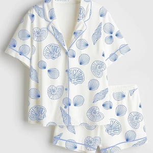 May include: White pajama set with a blue seashell pattern. The set includes a short-sleeved button-down shirt and matching shorts, both trimmed with blue piping. The seashell design is a repeating pattern.