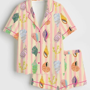 May include: A two-piece pajama set featuring a short-sleeved button-up shirt and matching shorts. The set has a light yellow and pink striped background with a seashell pattern in various colors. The edges are trimmed with pink piping.