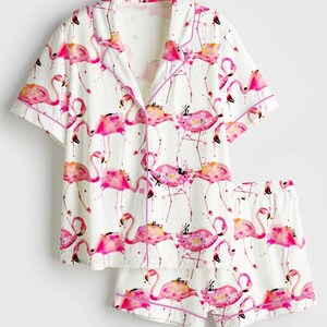 Op de afbeelding: Witte pyjamaset met een roze flamingo-print. De top met korte mouwen en de shorts hebben een herhalend patroon van roze flamingo's met aquarel-achtige details, afgewerkt met roze biezen.