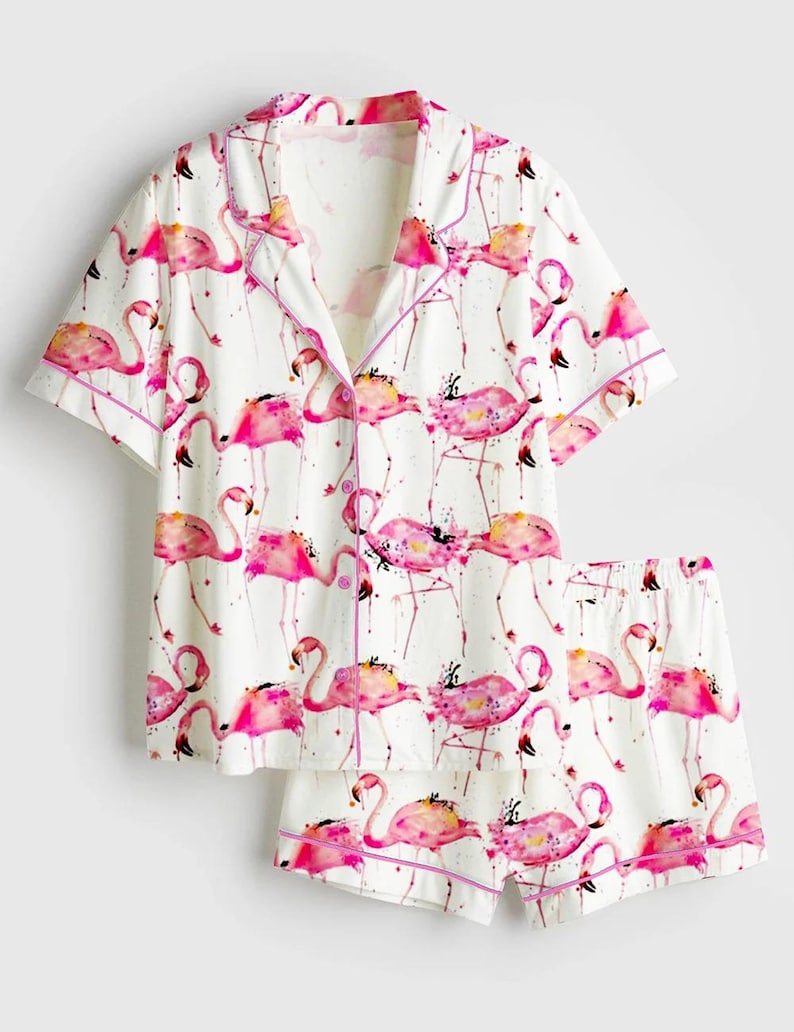 Op de afbeelding: Witte pyjamaset met een roze flamingo-print. De top met korte mouwen en de shorts hebben een herhalend patroon van roze flamingo's met aquarel-achtige details, afgewerkt met roze biezen.
