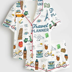 May include: White pajama set with a travel theme. The short-sleeved top and shorts feature a repeating pattern of travel icons, including bicycles, landmarks, and the text "Travel Planner". Red trim accents the edges.