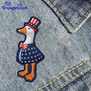 May include: An embroidered patch featuring a white goose wearing a red, white, and blue top hat, a red and white striped bow tie, and a blue dress with white stars. The goose has an orange beak and orange feet, set against a blue denim fabric.