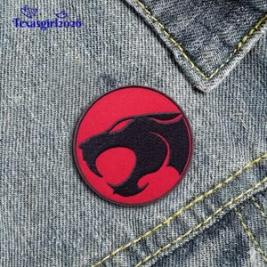 May include: A round, red patch featuring a black silhouette of a stylized cat-like creature. The patch is sewn onto a denim fabric background. The patch has a black border and is likely an iron-on or sew-on patch.