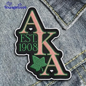 May include: Embroidered patch featuring the letters AKA in pink, outlined in black, with a green ivy leaf and the text "EST. 1908." The patch is on a denim background. The patch is a decorative item.