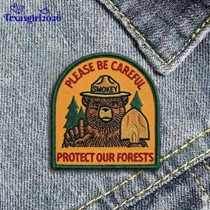 May include: Embroidered patch featuring Smokey Bear in a ranger hat, with the text "PLEASE BE CAREFUL" and "PROTECT OUR FORESTS". The patch has a green border and is set against a denim background.