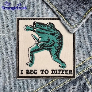 May include: Embroidered patch featuring a teal frog holding a knife, with the text "I BEG TO DIFFER" in black. The patch has a black border and is set against a cream background. The patch is on a denim background.