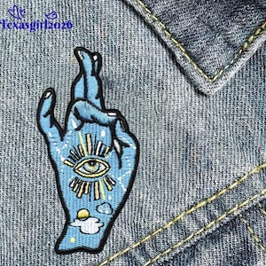 May include: A light blue embroidered patch in the shape of a hand with the fingers crossed. The patch features an eye design in the center, surrounded by rays and stars. The text "Texasgirl2020" is in the upper left corner. The patch is on a denim background.