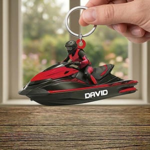 May include: A black and red jet ski keychain with a rider in a helmet and racing suit. The word "DAVID" is printed on the side of the jet ski. The keychain is held by a hand.