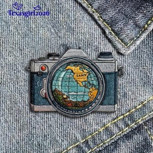 May include: Embroidered patch in the shape of a vintage camera with a globe lens. The globe shows North America in gold. The camera is blue and silver, with a red button. The patch is on a denim background.