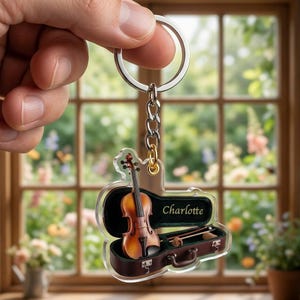 May include: A clear acrylic keychain featuring a miniature brown violin inside an open brown violin case with green lining. The name Charlotte is printed in gold script inside the case. The keychain includes a silver metal ring and chain.