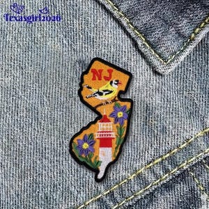 May include: Embroidered patch in the shape of New Jersey, featuring a yellow bird, a red and white lighthouse, and purple flowers. The patch has the letters "NJ" at the top and is set against a denim background.