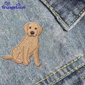 May include: An embroidered patch featuring a light brown Labrador retriever dog sitting in a profile position. The dog has dark brown eyes and a dark brown nose. The patch is placed on a blue denim fabric background with the text texasgirl2026.